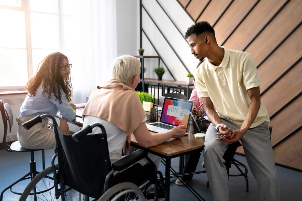 A collaborative office meeting where a diverse team of four professionals is gathered around a wooden desk. A woman in a wheelchair and another woman seated at the table look toward a man who is leaning against the desk, while a third man stands nearby. They are looking at a laptop screen in a bright, modern room with large windows and a decorative wood-paneled wall.