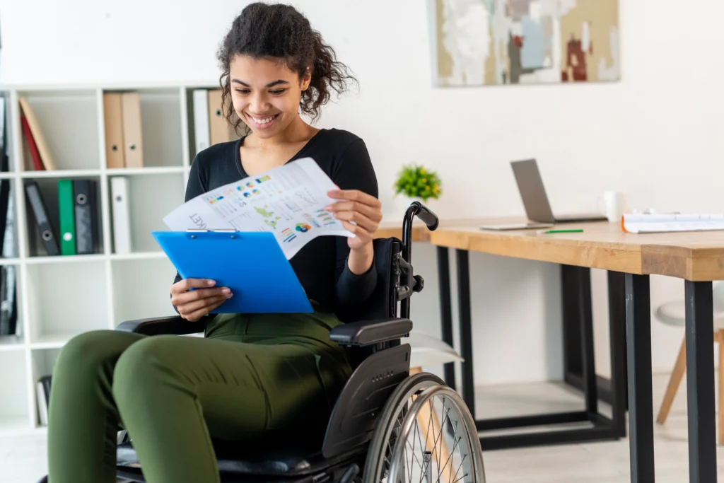 A diverse team of professionals sits around a table in a bright office discussing NDIS plan management support, which is often a key part of effective NDIS coordination in Adelaide and South Australia. One person in the foreground uses a wheelchair as the group collaborates near a whiteboard.