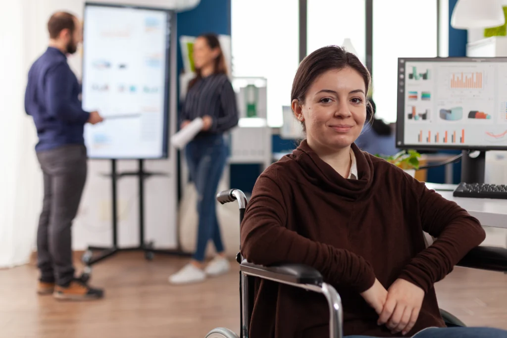 A woman in a brown sweater sits in a wheelchair in the foreground, smiling at the camera in a professional office environment. In the background, two colleagues are engaged in a presentation near a large screen displaying data charts, while a computer monitor on a nearby desk shows various colorful graphs and analytics.