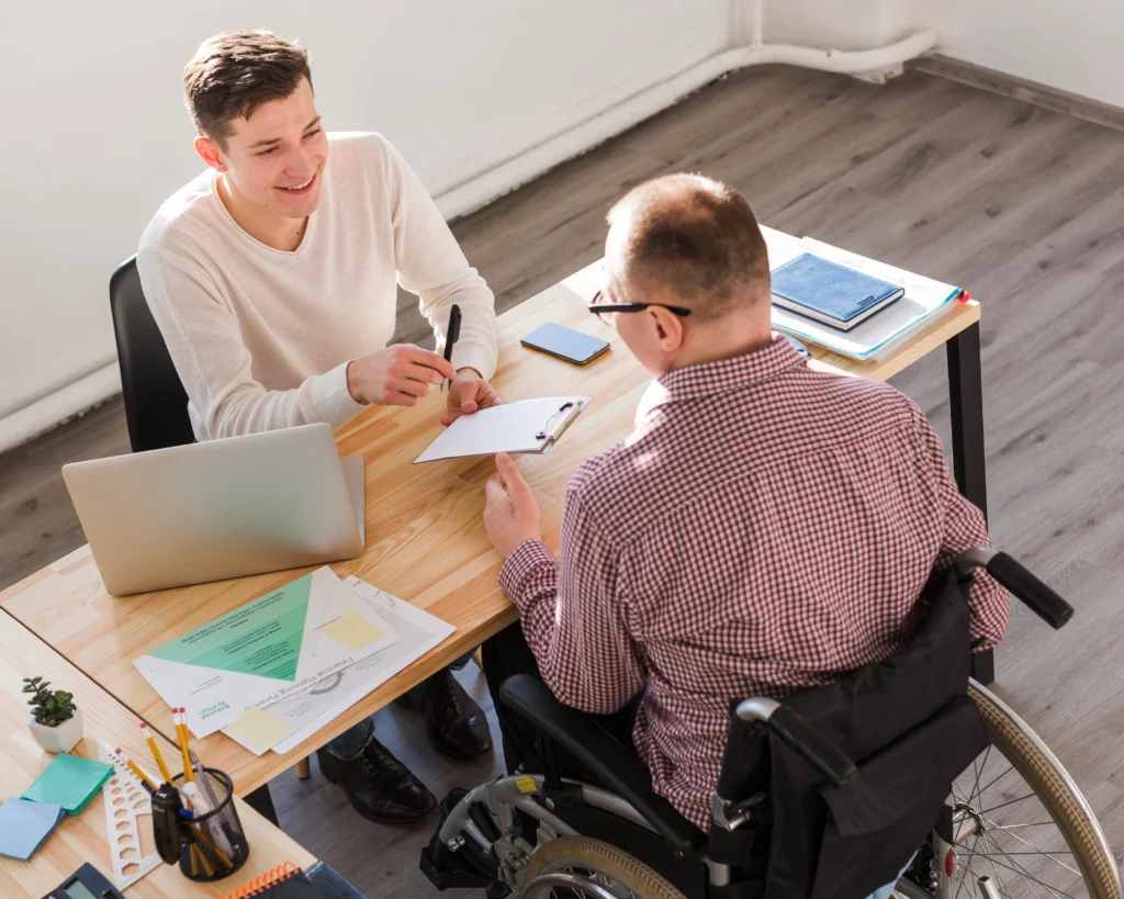 High-angle view of a professional meeting taking place at a light wood desk. A young man in a white sweater smiles while holding a pen and a clipboard, gesturing toward a man in a wheelchair who is seated opposite him. The desk is organized with a laptop, documents, and office supplies, highlighting a supportive planning session.