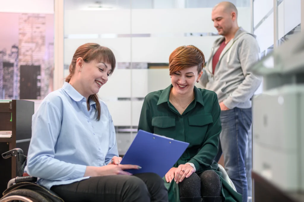 Two women, one of whom is using a wheelchair, are smiling and looking at a blue clipboard together in a bright office setting. In the blurred background, a man stands near a glass partition. The scene conveys a positive, supportive interaction between a professional and a client.