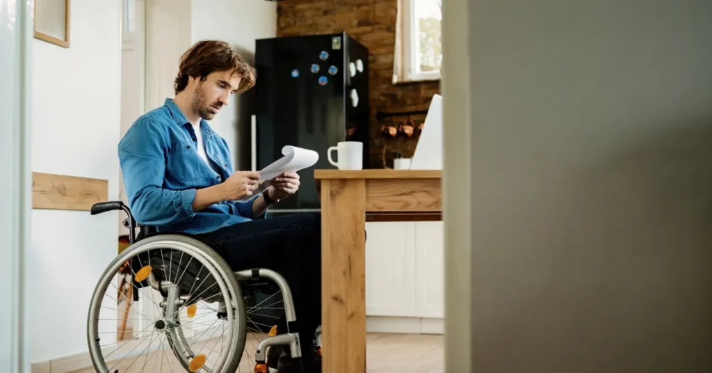 Man using a wheelchair sitting at a dining table, reading papers in a bright, accessible home kitchen setting.